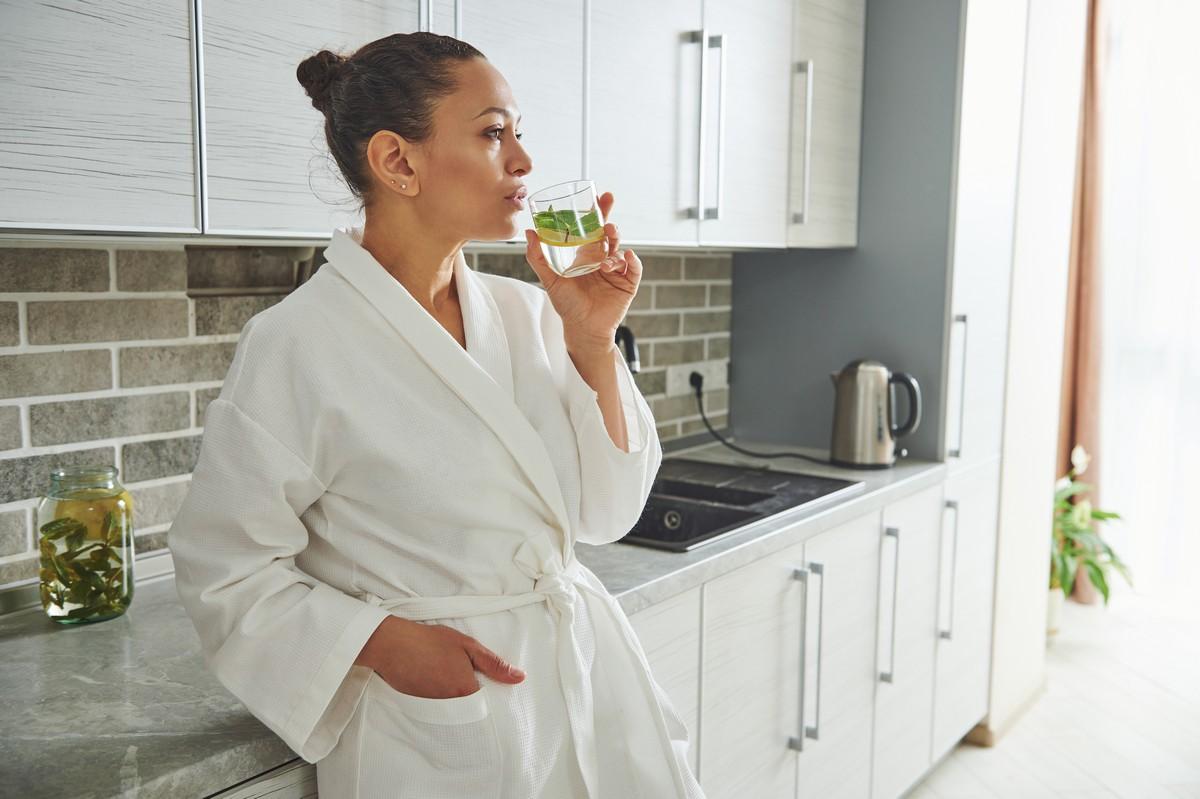 A lady in a white dressing gown sips lemon water whilst leaning on her kitchen worktop.