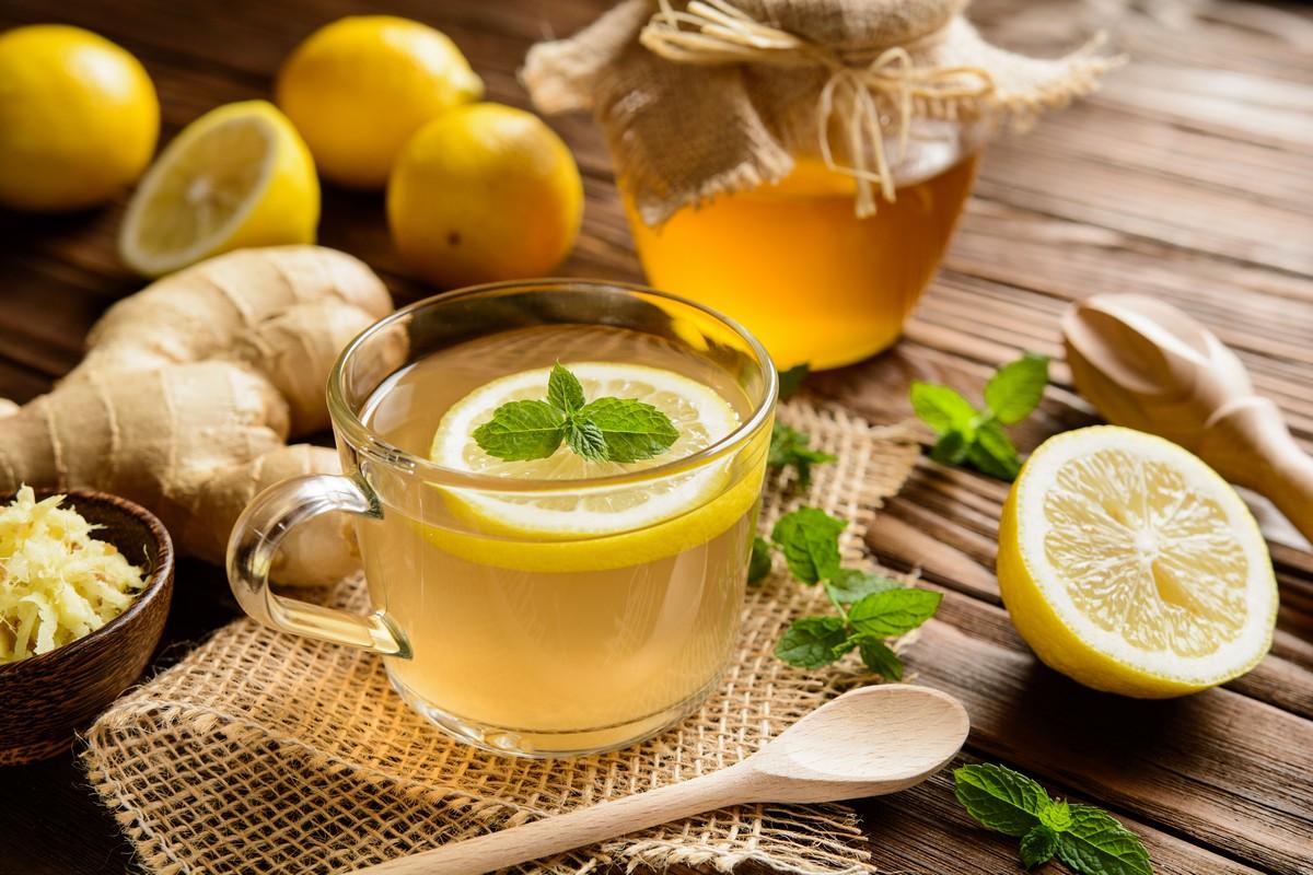 A clear mug of tea with a lemon wedge and mint leaves on top sits on a rustic wooden table. Next to it are whole lemons, a small bowl of grated ginger, a jar of honey, a wooden spoon, and scattered mint leaves.