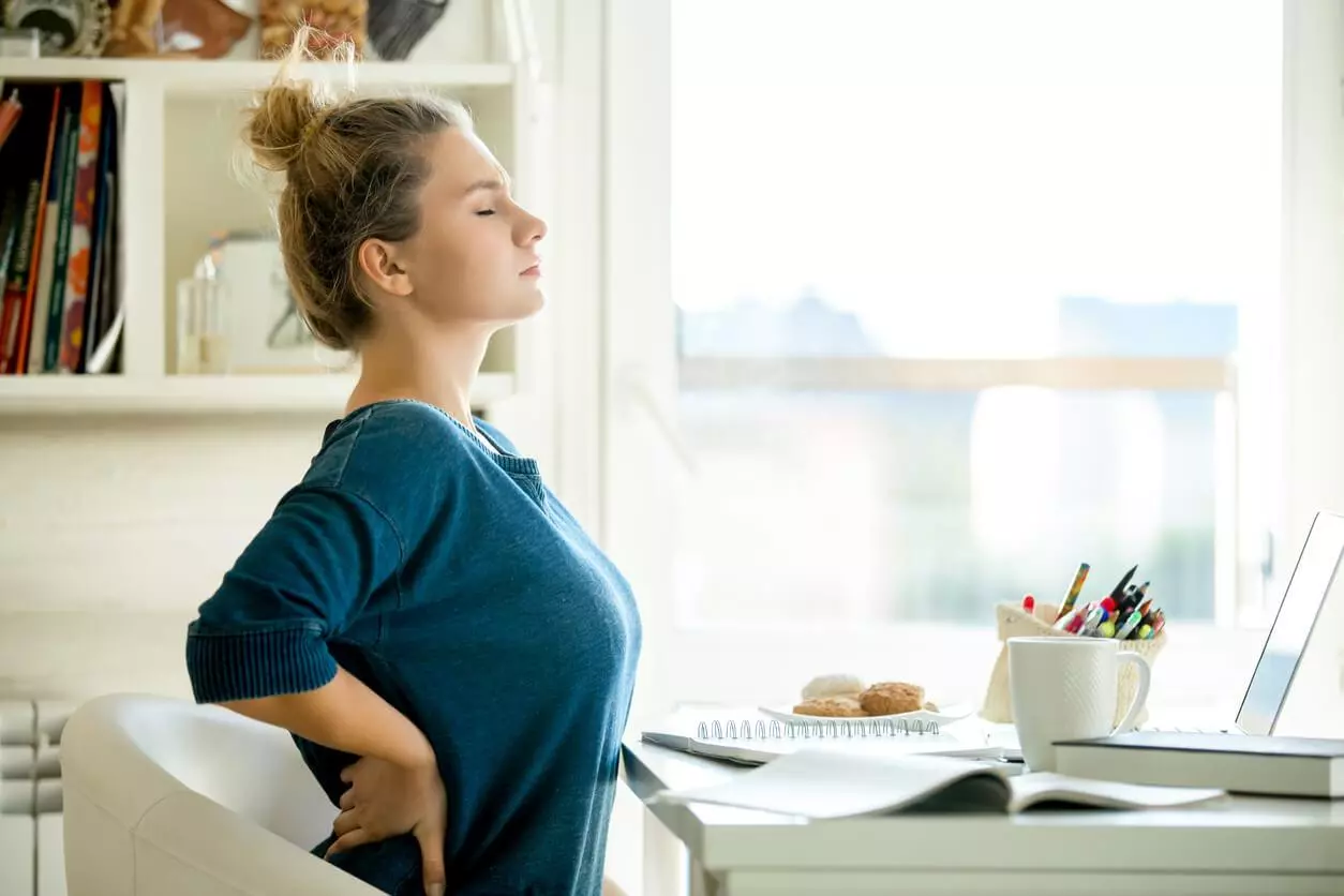 Image of a woman stretching her back by leaning backwards while sitting at a table, with her eyes closed and a hand on her lower back, suggesting relief from back pain.