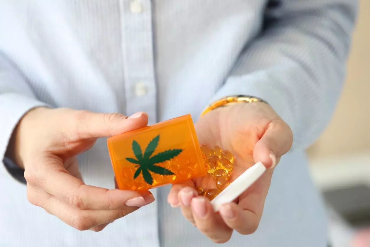 A person's hands holding an orange prescription bottle with a cannabis leaf symbol, spilling out clear, amber-coloured gel capsules into their palm, suggesting medical marijuana pills for therapeutic use.
