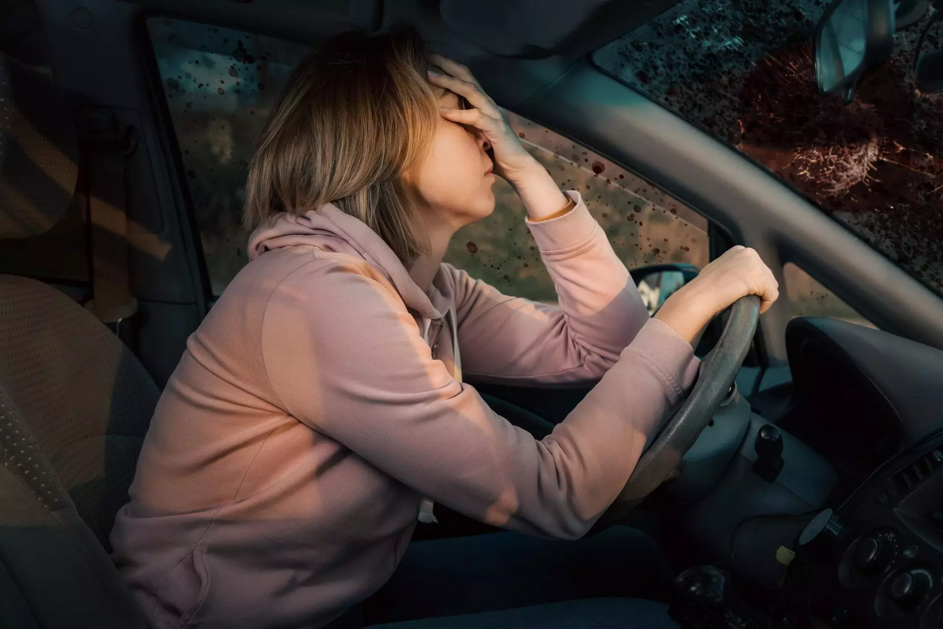 A woman seated in the driver's seat of a car with the steering wheel on the left side, shielding her eyes with her hand.