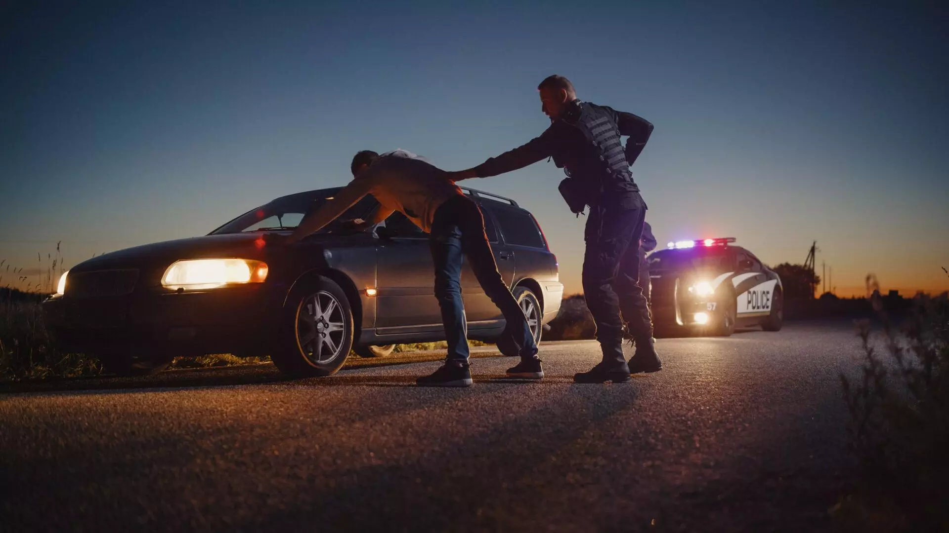 A law enforcement officer performing a pat-down on a driver who is standing with his hands on the car's hood.