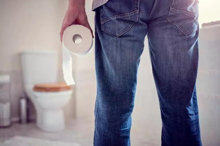 A man in a bathroom holds a roll of toilet paper, standing next to a toilet, emphasising a common household situation.