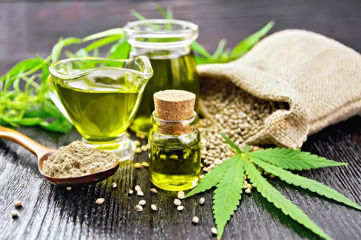 A vibrant display of cannabis-derived products, including green cannabis oil in glass bottles, a spoonful of cannabis powder, and raw hemp seeds, accentuated by fresh cannabis leaves.