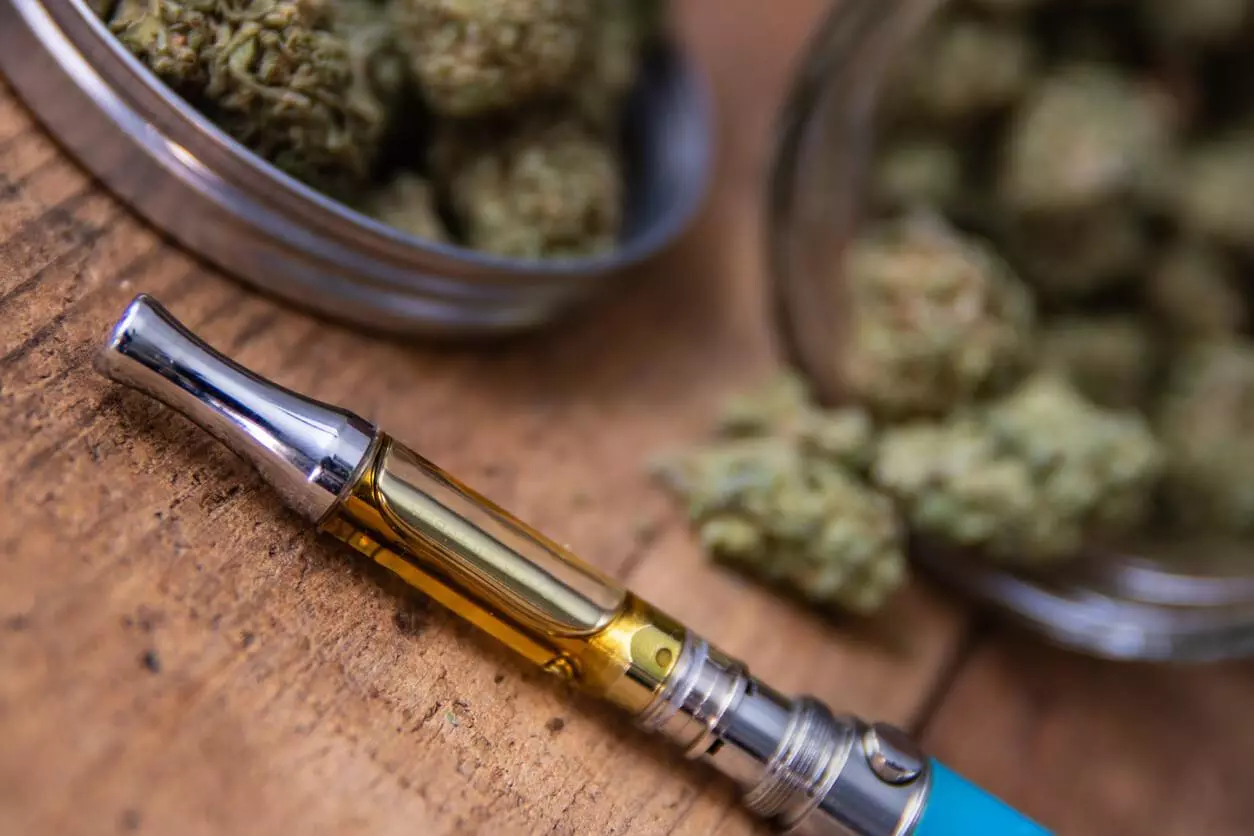 A close-up image of a cannabis oil vape pen alongside a container full of cannabis buds.