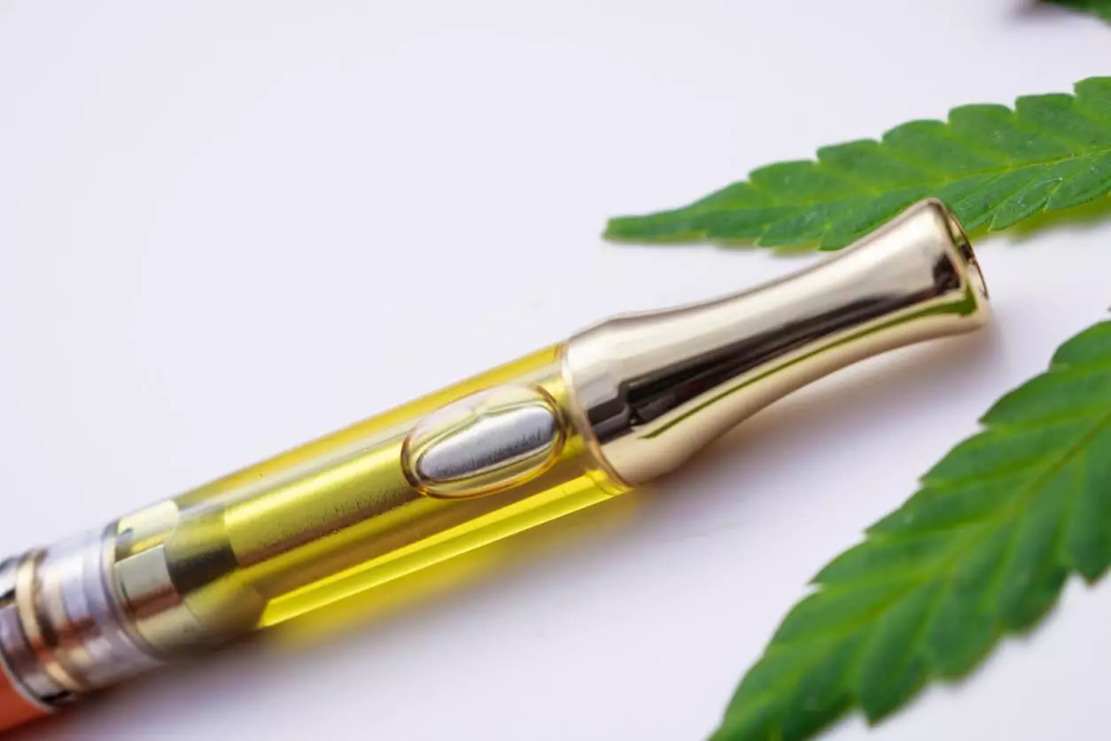 A close-up image of a cannabis oil vape pen with a golden hue, lying next to a cannabis leaf on a white background.