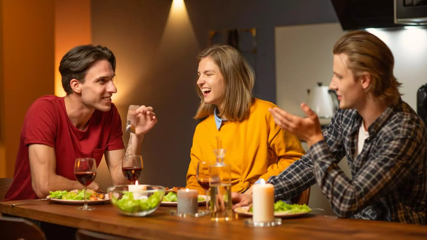 Three friends enjoy a lively conversation over dinner with wine in a cosy, warmly lit home setting. 