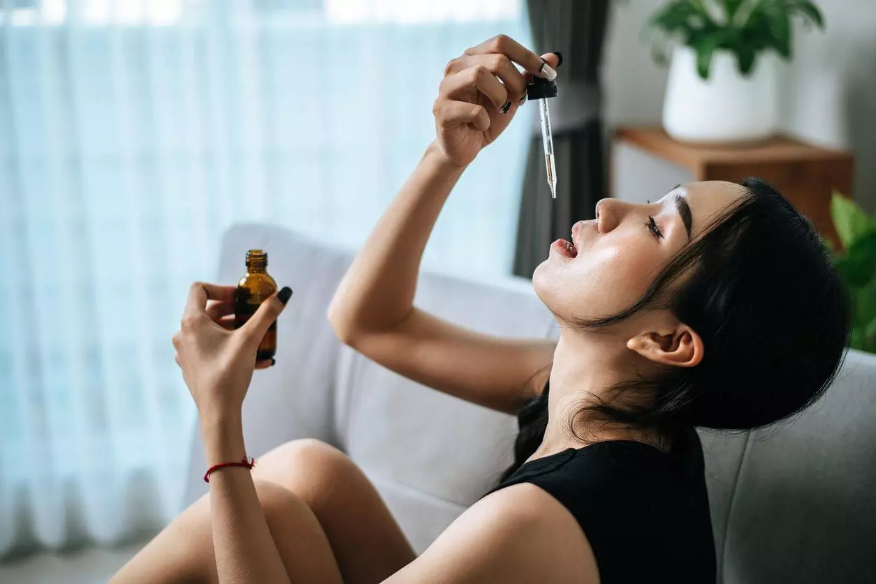 A woman sitting on a couch using a dropper to take cannabis oil sublingually.