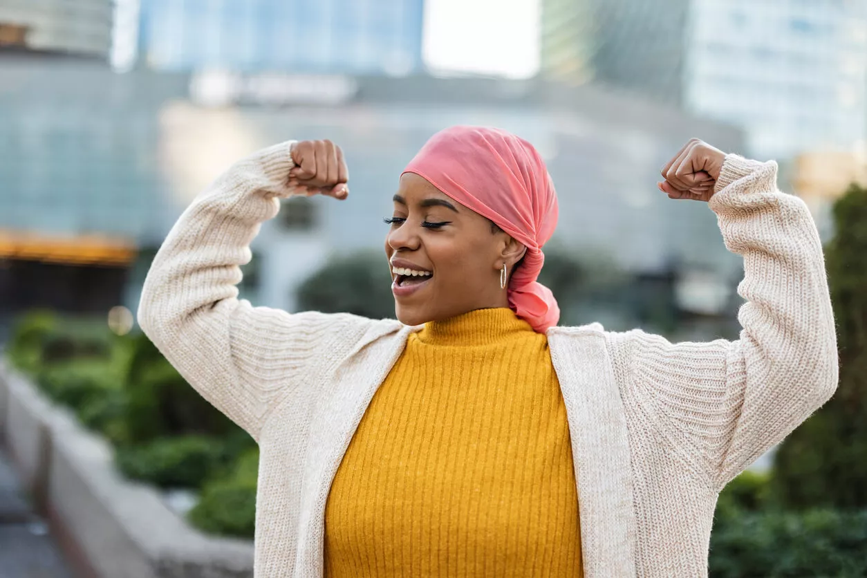 A woman stands outdoors, smiling and flexing her arms in a strong pose. She is wearing a pink headscarf, a mustard yellow sweater, and a cream-coloured cardigan.