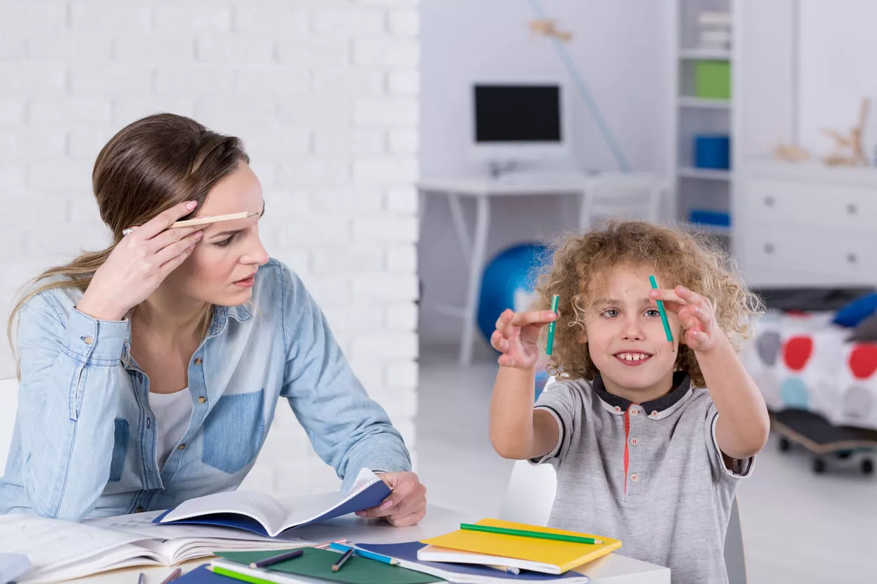 A frustrated woman looks at a book while a smiling, curly-haired child plays with two pencils. They sit at a table with notebooks and stationery in a bright room.