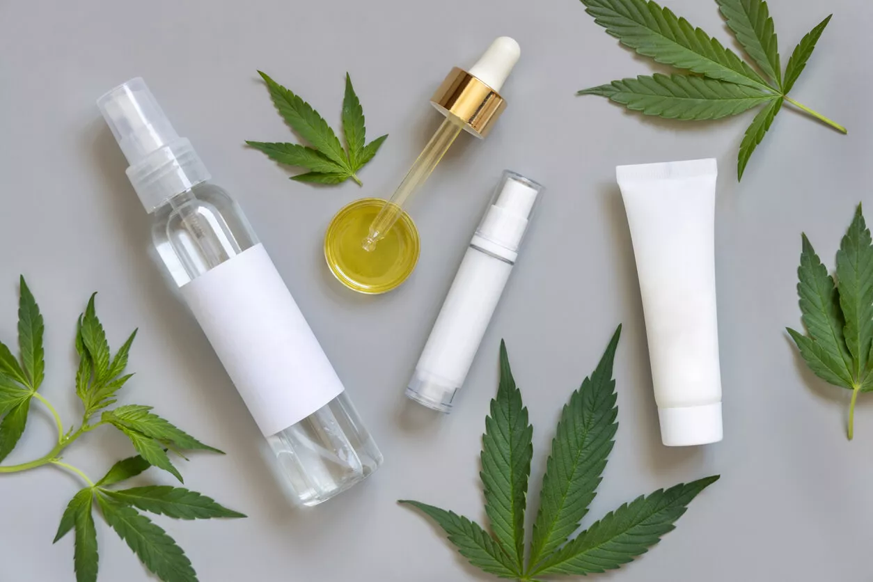 Various cannabis-based topical products, including a spray bottle, dropper with oil, and cream tubes, are arranged on a grey surface, surrounded by cannabis leaves.